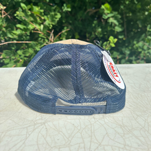 Load image into Gallery viewer, A blue mesh-back trucker cap with a tan fabric front panel and a white hangtag, photographed outdoors on a light-colored surface with green foliage in the background.
