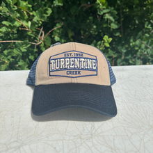 Load image into Gallery viewer, A beige and navy blue trucker hat with 'Turpendine Creek' embroidered on the front, resting on a light-colored textured surface with a background of green foliage.
